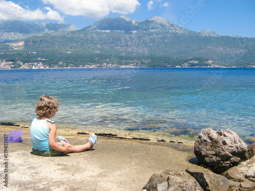 Cute curly toddler girl with butterfly net for catching fish sitting on the seashore, lookig at sea