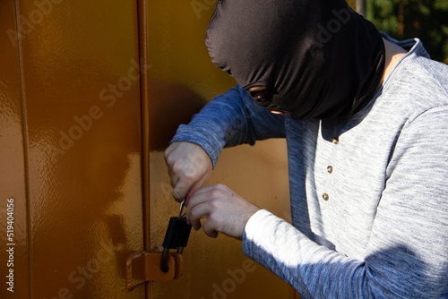 a thief in a black mask picks open a padlock on a metal door