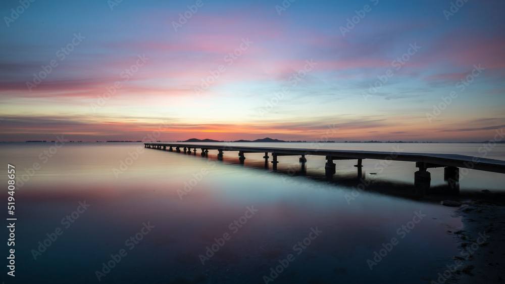 Beautiful wide landscape at sunrise in the Mar Menor, Cartagena, Spain. With a spectacular  very colorful sky and reflected in the calm waters of the sea. We also see a wooden jetty on the water. It t