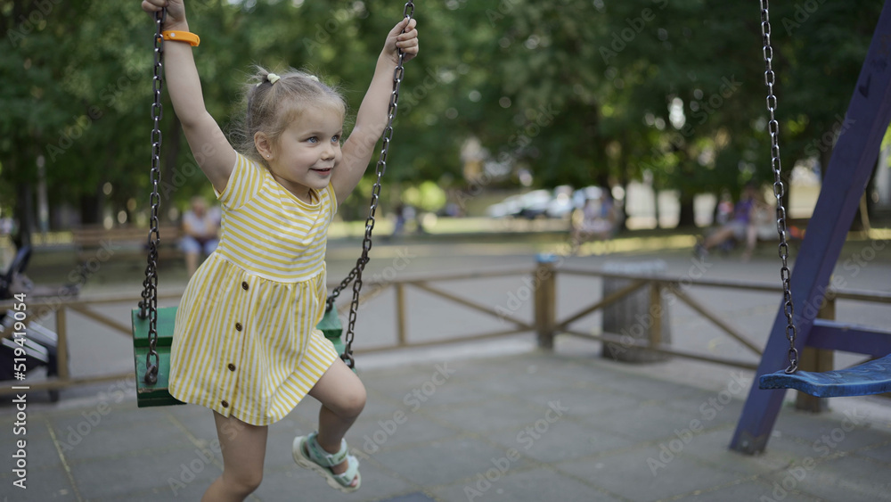 Cute little girl rides on a swing in the playground. Child girl playing on the playground in the city park Cute little girl rides on a swing in the playground. Child girl playing on the playground in the city park