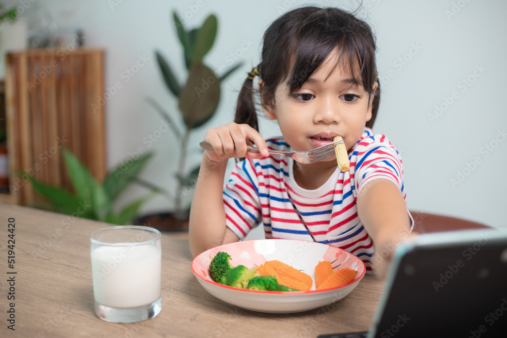 Adorable Asia child girl having lunch while watching a movie from the tablet. A little Asian child eating dinner and eyes are looking cartoon from Tablet. National Eating Disorders Awareness Week. Adorable Asia child girl having lunch while watching a movie from the tablet. A little Asian child eating dinner and eyes are looking cartoon from Tablet. National Eating Disorders Awareness Week.