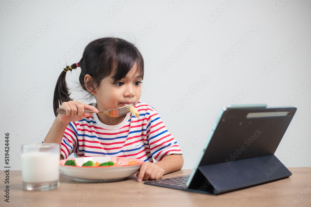 Adorable Asia child girl having lunch while watching a movie from the tablet. A little Asian child eating dinner and eyes are looking cartoon from Tablet. National Eating Disorders Awareness Week. Adorable Asia child girl having lunch while watching a movie from the tablet. A little Asian child eating dinner and eyes are looking cartoon from Tablet. National Eating Disorders Awareness Week.