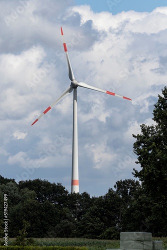 A portrait of a big windmill standing tall above the trees with a cloudy sky behind it. The turbine generates green clean ecological sustainable and electric energy by catching wind on its wings.