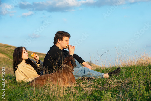young loving couple on a picnic in nature with their pet, a big red dog against a bright blue sky. Young people eating fast food outdoors