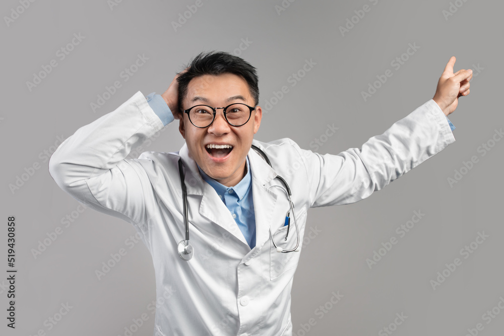 Shocked excited adult asian male doctor in white coat, glasses with open mouth pointing finger at empty space