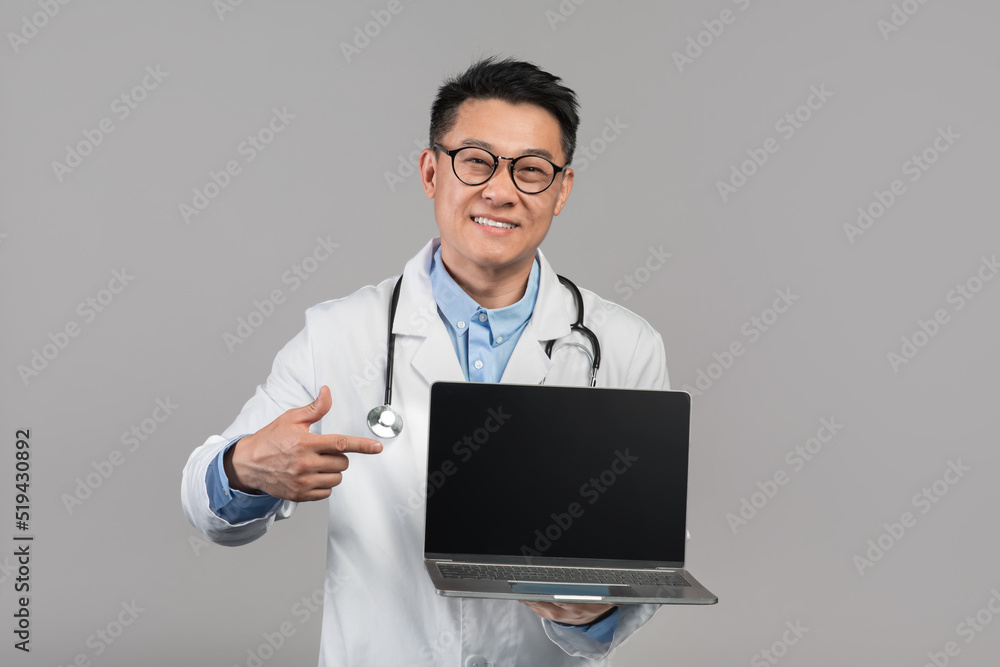Smiling mature korean male therapist in white coat in glasses, show finger on laptop with blank screen Smiling mature korean male therapist in white coat in glasses, show finger on laptop with blank screen