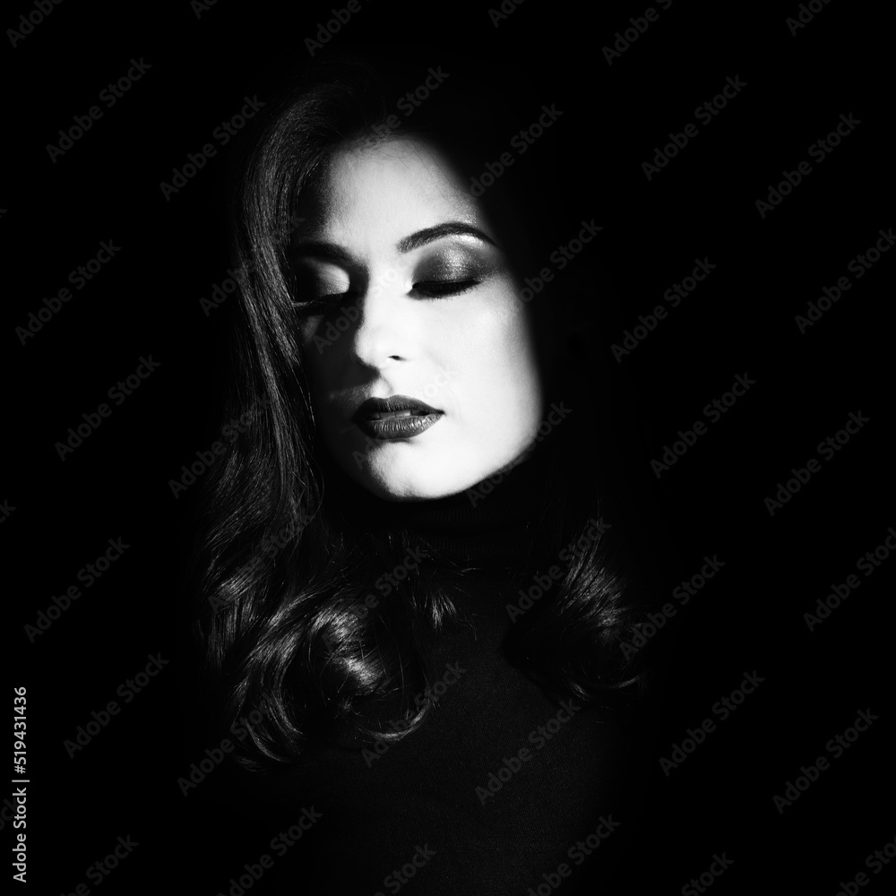 Beauty and fashion concept. Beautiful woman portrait standing in darkness with illuminated face. Model with evening makeup and wavy hair with closed eyes. Black and white image