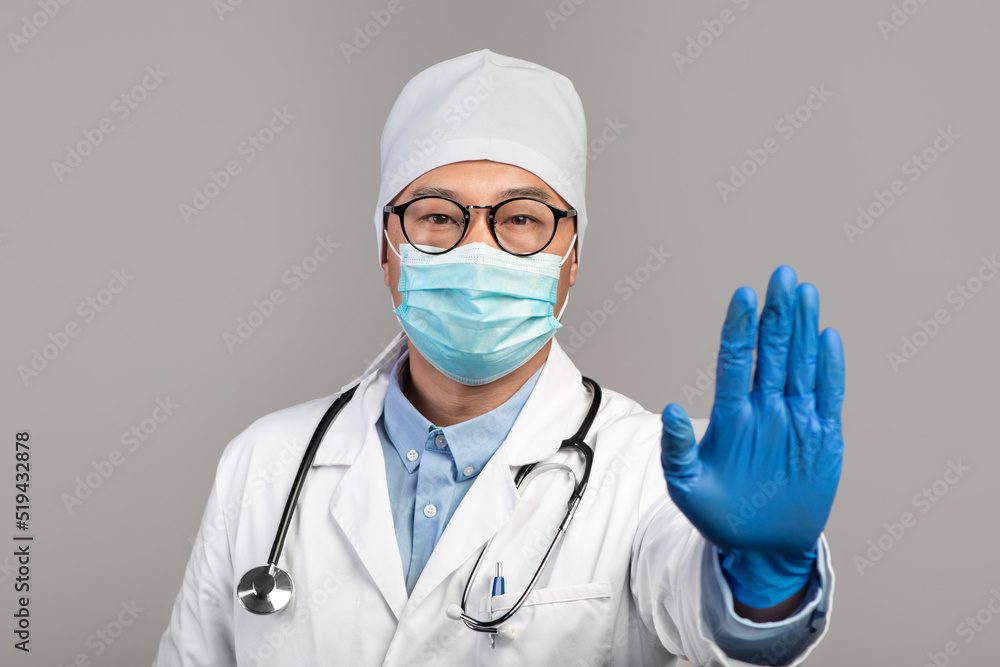 Serious adult asian doctor in white coat, glasses and protective gloves shows stop sign with hand Serious adult asian doctor in white coat, glasses and protective gloves shows stop sign with hand