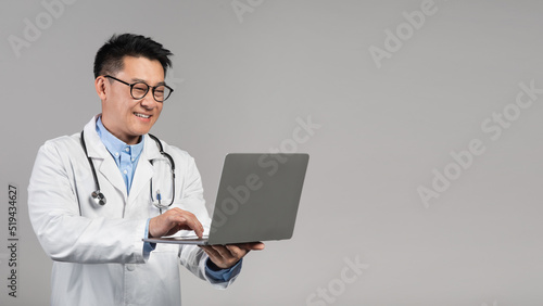 Glad smiling confident middle aged asian doctor in white coat, glasses with stethoscope typing on laptop
