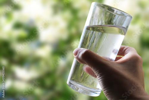 A glass of water in a hand close-up on a natural green background outdoors.