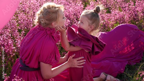 fashionable beautiful woman in a long pink dress with a child daughter girl lying on a field with pink flowers on the field in summer .