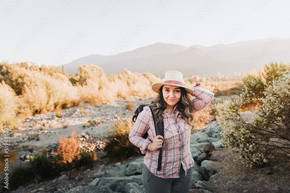 Smiling young latin woman with a hat, a pale pink blouse and a backpack on, hiking in a beautiful landscape.