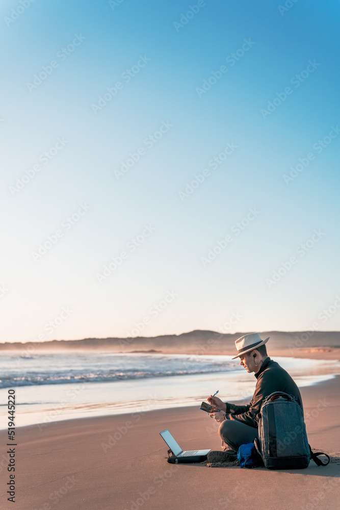 Fototapeta premium digital nomad sitting on the beach with his laptop and taking notes diary on the shore of the beach at sunset
