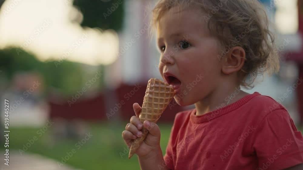 Fair-haired boy eats white cold cream ice cream in waffle cone cup while sitting outside in sunny weather at sunset time. High quality 4k footage Fair-haired boy eats white cold cream ice cream in waffle cone cup while sitting outside in sunny weather at sunset time. High quality 4k footage