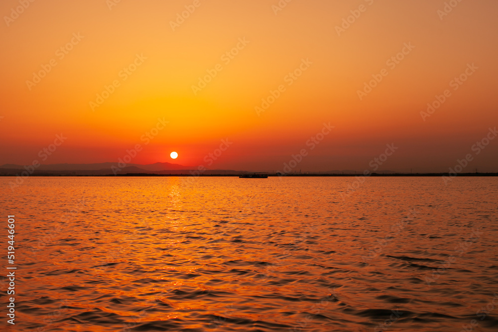 Fototapeta premium Colorful orange, yellow and red sunset over the water of lake with sun setting in the water. Natural park at sunset with distant silhouettes of mountains. Sunset landscape view from a boat. Traveling 