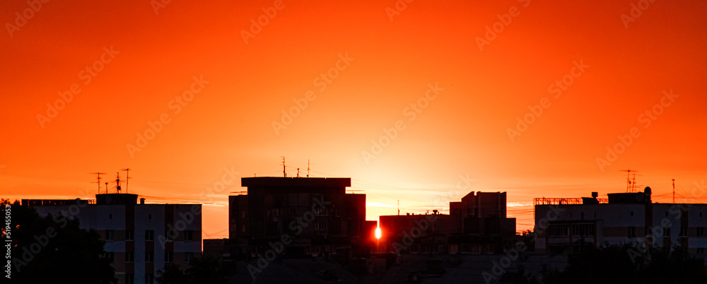 Silhouette of city at sunset. Sun is rising over tall buildings. Dramatic sky in sun rays. Urban landscape at dawn. Copy space, banner format.