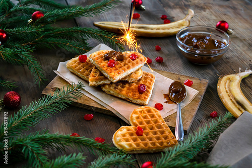 Homemade Belgian waffles heart shaped on plate with colorful red sweet cherry berries, banana, and boiled condensed milk,flaming bengal fire. Christmas new year background,dessert food