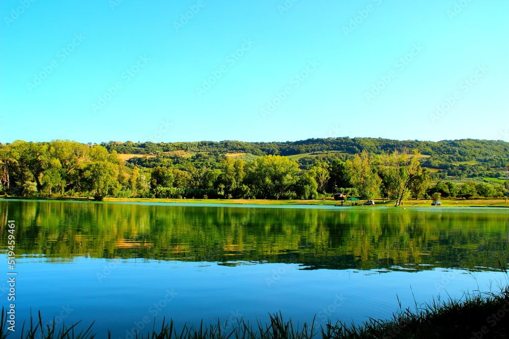 Clear view from Lago Cedri in Lapedona with the balmy surface of gentle water extending over the picture and reflecting the various vegetation in it while being compounded by the splendid Marche hills
