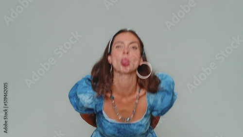 Cheerful funny bully young woman in dress. Adult girl showing tongue making faces at camera, fooling around, joking, aping with silly face, teasing isolated alone on gray studio background indoors