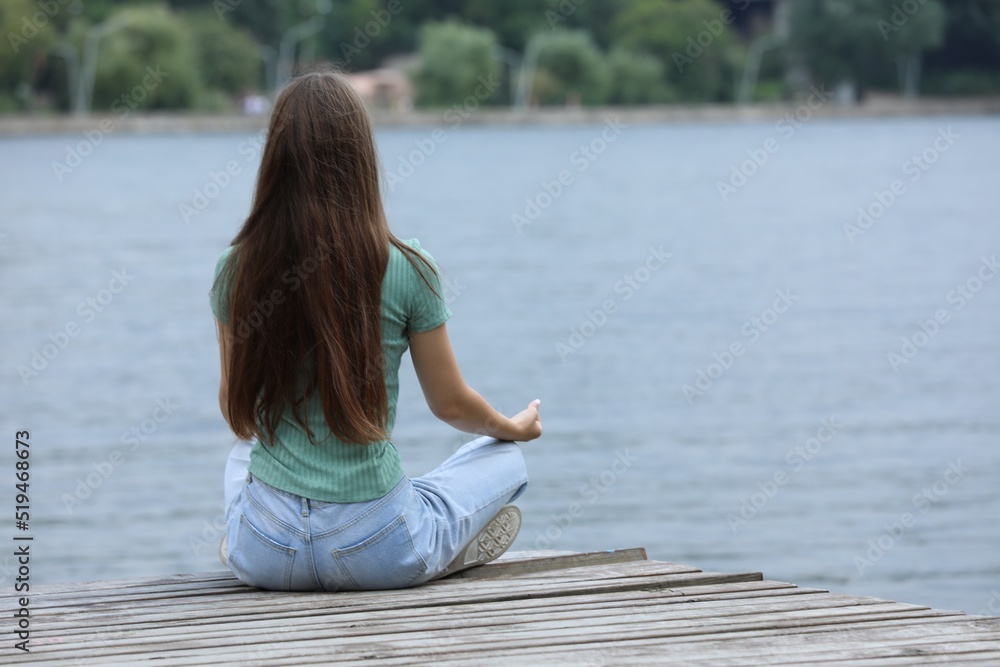 Teenage girl meditating near river, back view. Space for text