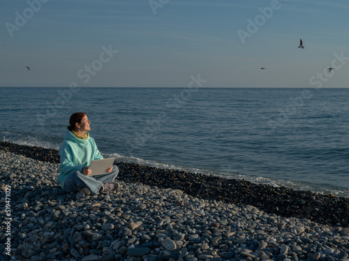 Happy caucasian woman working on a laptop while sitting on a pebble beach. © Михаил Решетников