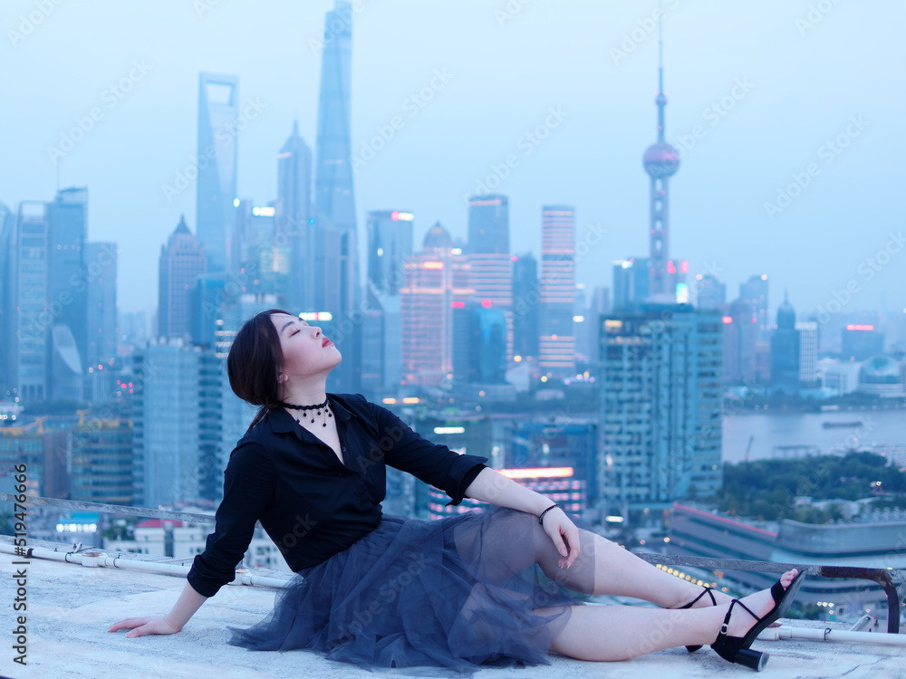 Beautiful young Chinese woman in black sitting on top of mansion roof with blur Shanghai Bund landmark buildings background with dusk light. Emotions, people, beauty and lifestyle concept. Beautiful young Chinese woman in black sitting on top of mansion roof with blur Shanghai Bund landmark buildings background with dusk light. Emotions, people, beauty and lifestyle concept.