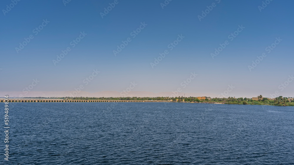 The dam of the sluice system in Esna. A long concrete bridge crosses a wide river. Green vegetation is visible far away on the shore. Clear blue sky. Copy space. Egypt. Nile