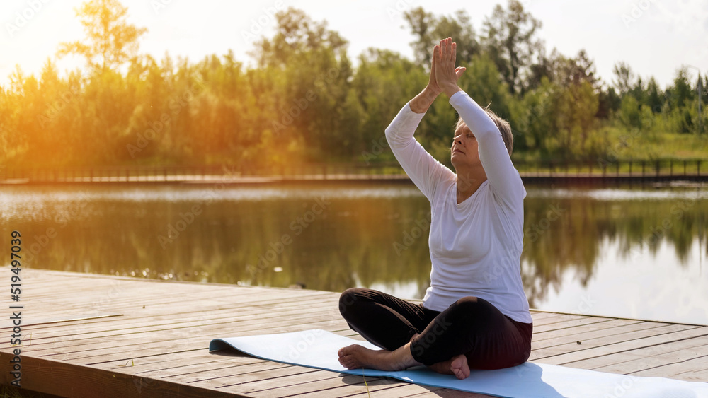 Caucasian mature woman in a sports outfit, relaxing, meditating, on a fitness mat in a public outdoor park. Healthy active lifestyle of the elderly.