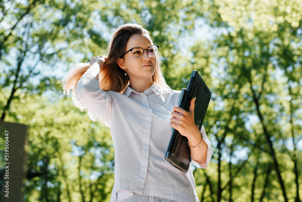 Female student wearing eyeglasses holding laptop and smartphone touching her hair while standing in park and looking away Female student wearing eyeglasses holding laptop and smartphone touching her hair while standing in park and looking away