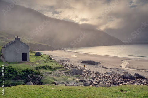 Calm and moody nature scene with small house tourist camp by a beach and mountains. Low cloudy sky and fog. Keem bay area, county Mayo, Ireland. Famous travel are for water sport and hiking.