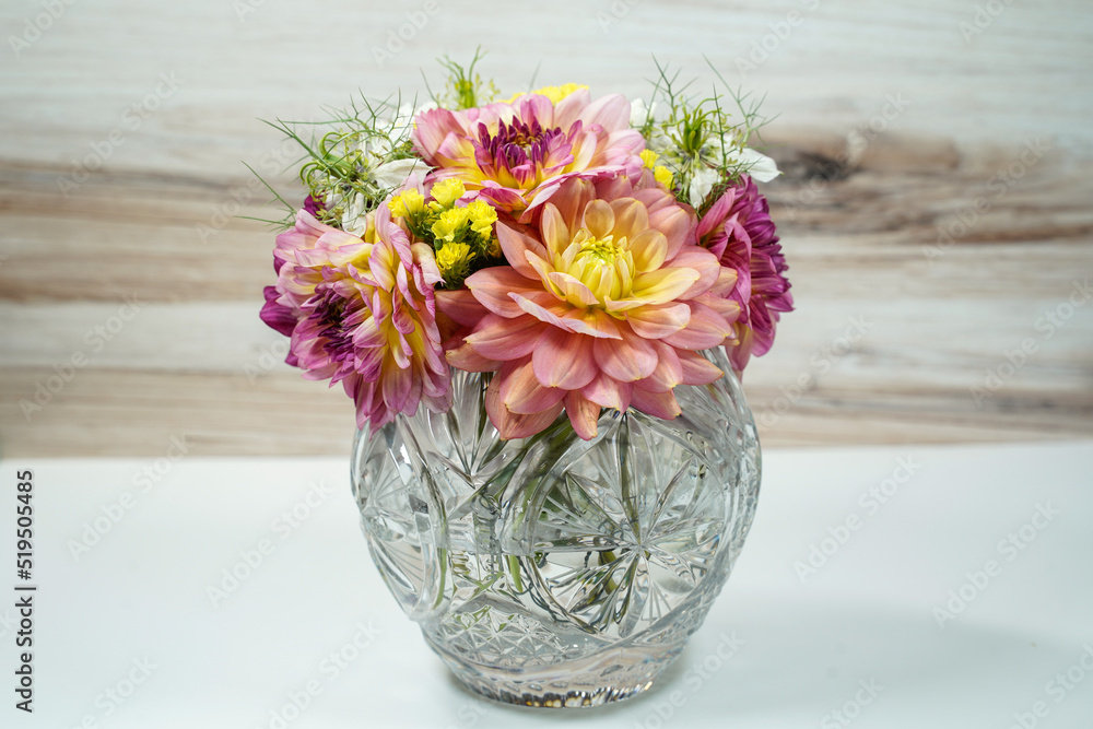 Round glass bowl filled with a bouquet of dahlia and statice flowers. Pink, purple, yellow, and white flowers. White table with wood background. Round glass bowl filled with a bouquet of dahlia and statice flowers. Pink, purple, yellow, and white flowers. White table with wood background.