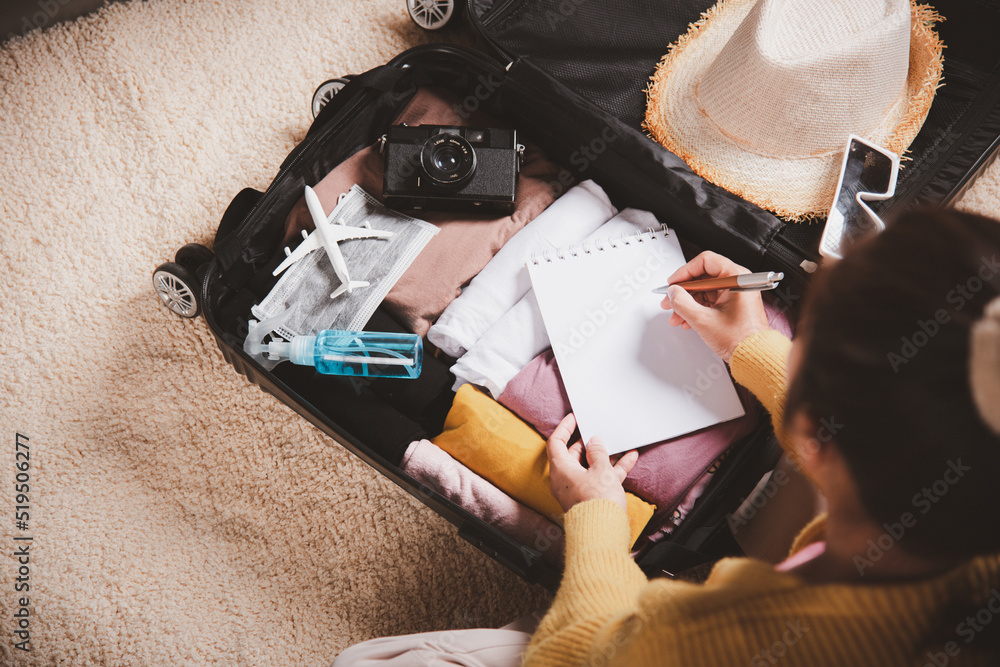 Making check list of things to pack for travel. Woman writing paper take note and packing suitcase to vacation writing paper list sitting on room, prepare clothes into luggage, Travel vacation travel - obrazy, fototapety, plakaty 