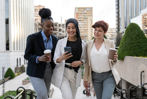 three multiracial female businesswomen of different ages walking outdoors with tablet and coffee, leaving office and meeting outdoors photo