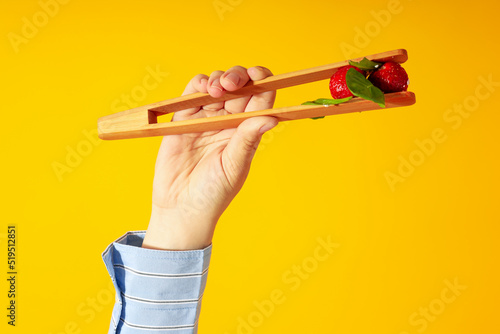 Female hand holds salad with strawberry with wooden tongs on yellow background