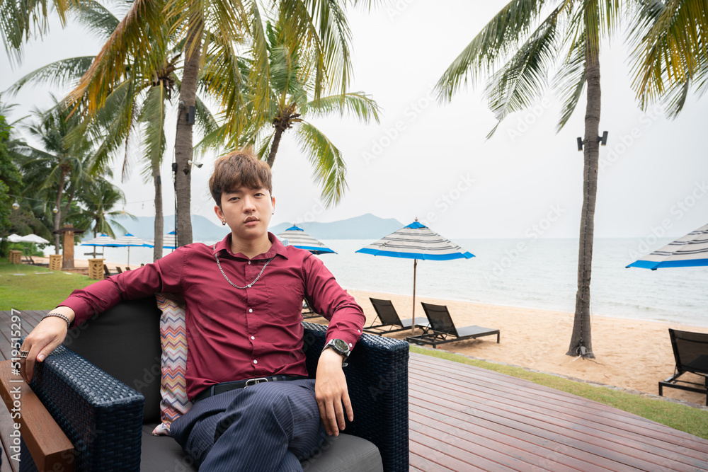 Fototapeta premium Portrait of handsome asian man red shirt siting on chair and posting over beach background. 