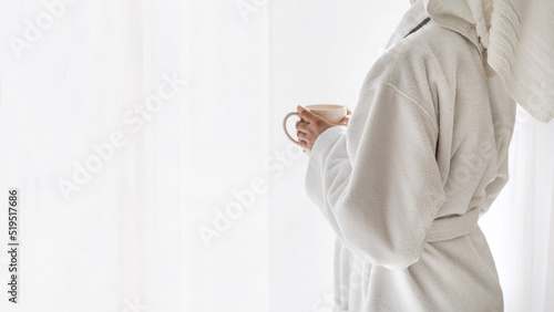 Woman in bathrobe standing near window with coffee cup