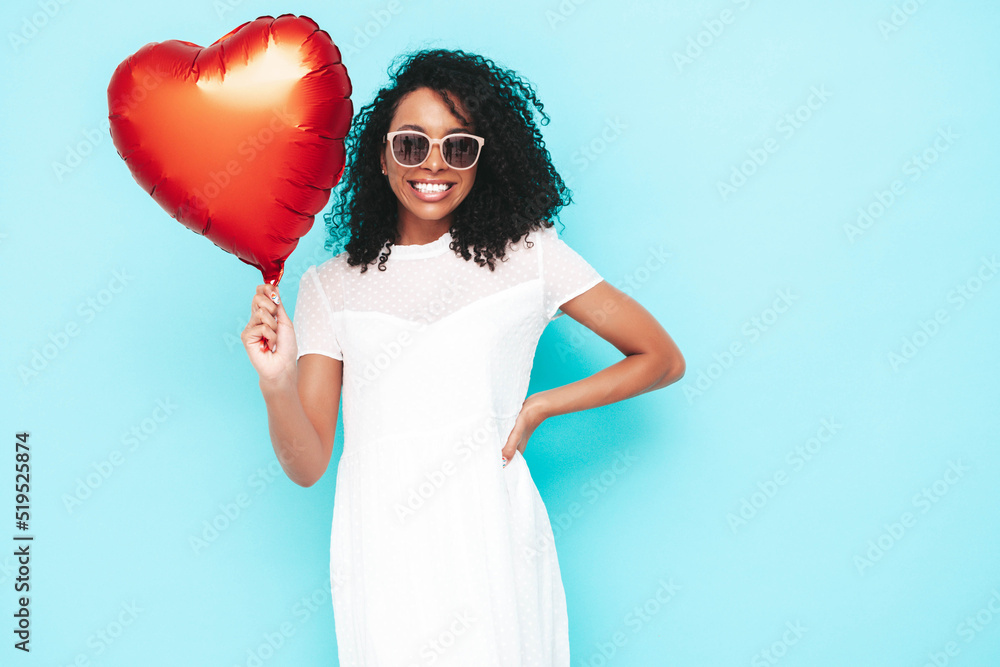 Beautiful black woman with afro curls hairstyle. Smiling model dressed in white summer dress. Sexy carefree female posing near blue wall in studio. Tanned and cheerful. Holding heart air balloon Beautiful black woman with afro curls hairstyle. Smiling model dressed in white summer dress. Sexy carefree female posing near blue wall in studio. Tanned and cheerful. Holding heart air balloon