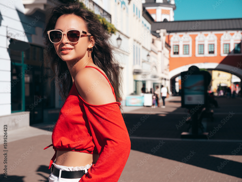 Young beautiful smiling hipster woman in trendy summer red top and shorts clothes. Sexy carefree model posing in the street at sunset. Positive brunette female. Cheerful and happy. In sunglasses Young beautiful smiling hipster woman in trendy summer red top and shorts clothes. Sexy carefree model posing in the street at sunset. Positive brunette female. Cheerful and happy. In sunglasses