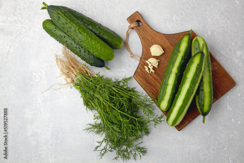Peeled fresh cucumbers on a brown wooden cutting board with a bunch of dill and garlic on a light gray concrete table