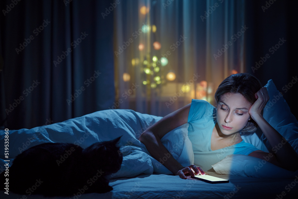 Young woman with her cat using smartphone in bed in her cozy bedroom at night. Young woman with her cat using smartphone in bed in her cozy bedroom at night.