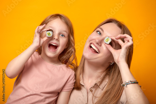 Funny mom with daughter holding sushi rolls in front of eyes on yellow background
