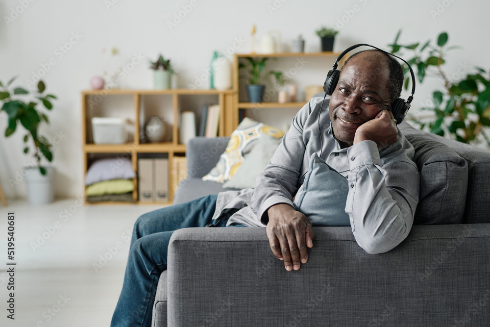 Portrait of African senior man in headphones smiling at camera while resting on sofa in living room and enjoying music