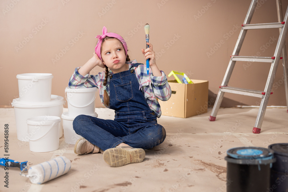 Naklejka premium A sad tired girl props elbow on the paint cans sighs lets the air out of mouth with an expression of lack of strength, holds in hand a paintbrush soiled with the brown color of the walls.