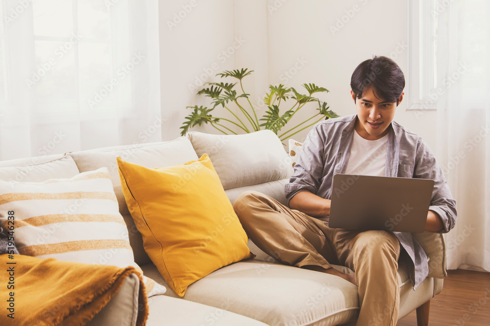 Handsome asian man t-shirt sits lobby sofa in the living room of his home connected to reading Internet messages. Look at your laptop computer screen cozy little corner of your home. Handsome asian man t-shirt sits lobby sofa in the living room of his home connected to reading Internet messages. Look at your laptop computer screen cozy little corner of your home.