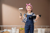 Cute little girl in pigtails dressed in a denim outfit and a pink bandama in her hair holds a paint tray and roller, apartment renovation concept, painting walls.