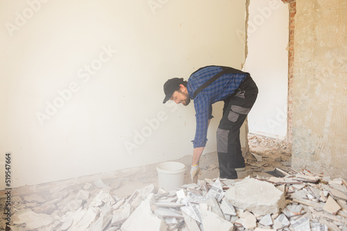 Fotografija A man in a work suit with a baseball cap cleans up in an apartment under renovation, picks up debris from the floor left after demolishing walls with a shovel