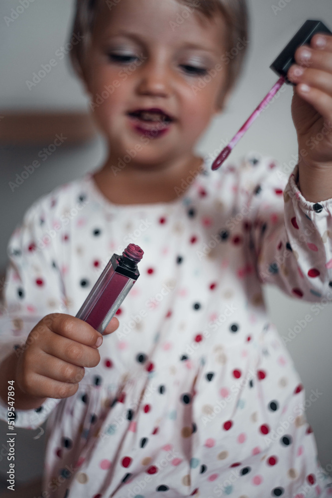 A little girl in a pink dress with short hair is playing with her mother's makeup. Smiling child