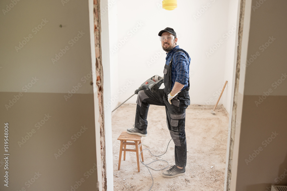 Smiling satisfied with work, a worker in work pants and shirt and safety goggles leans leg against a stool holds an impact hammer with a drill is repairing a house.