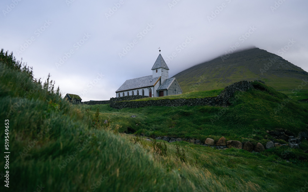 View on to the church of Vidareidi also named Vidareidi Kirkja with moody cloudy sky and mountains. Faroe Islands, Denmark, Europe.