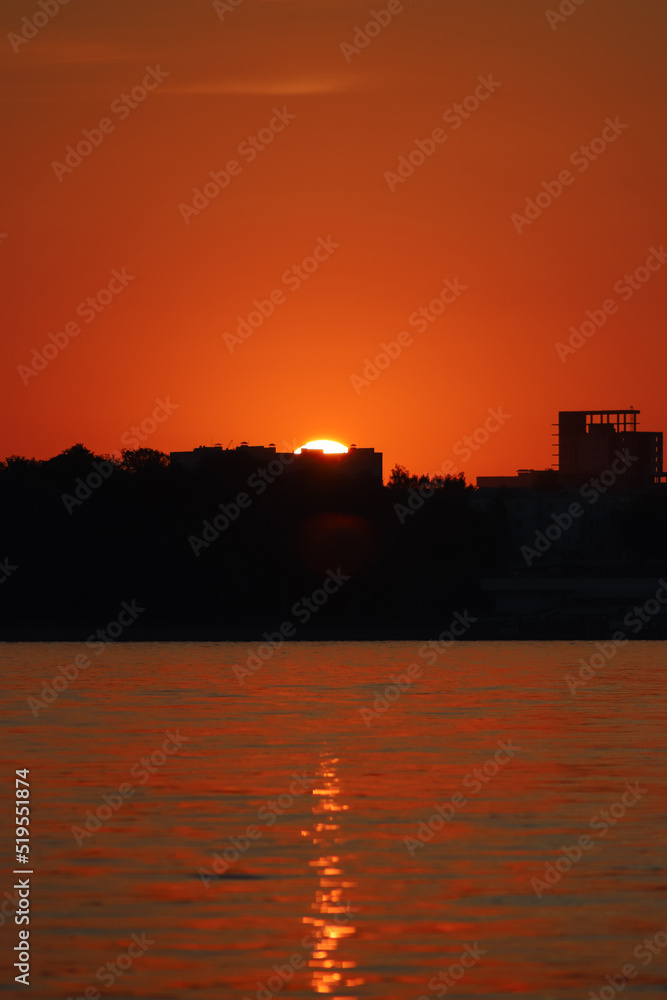 Fototapeta premium A bright setting sun sets behind the black silhouettes of buildings above the river against an orange red sky. Summer sunset background. Beautiful landscape, close up